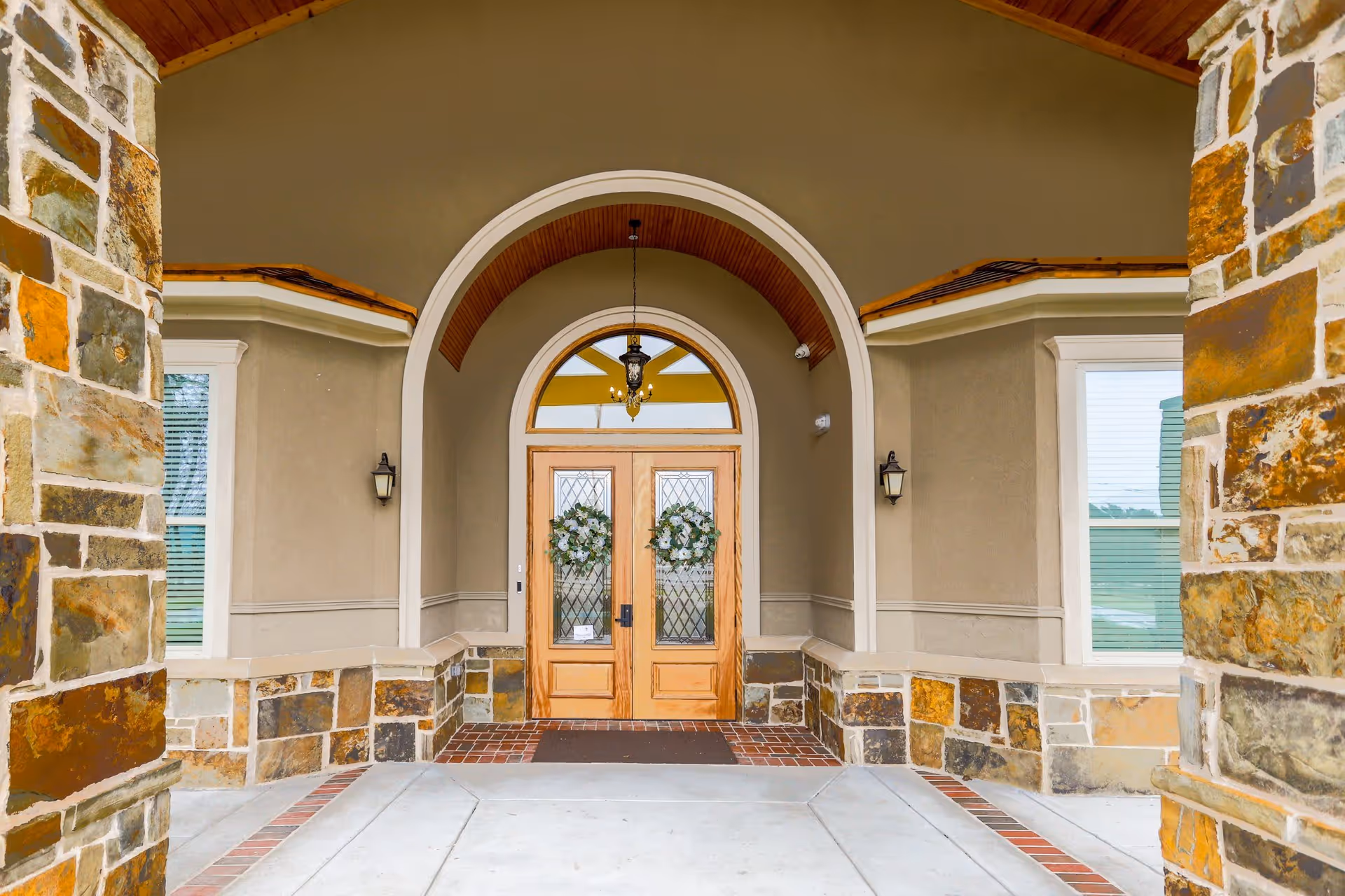 Entrance of a building with double wooden doors decorated with green wreaths, stone pillars on either side, and an arched ceiling with a hanging lantern light fixture.