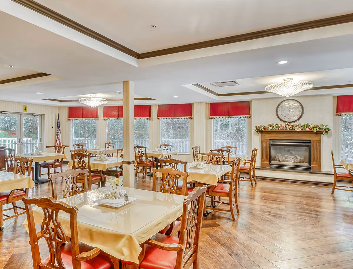 Bright dining room with multiple tables and chairs, a fireplace with a clock above, chandeliers, and large windows with red valances.