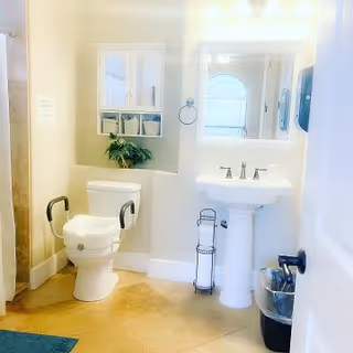 A bright and clean bathroom featuring a toilet with safety grab bars, a white pedestal sink with a mirror above it, a small cabinet with baskets mounted on the wall, a towel ring, a trash can, and a toilet paper holder. The floor is tiled and there is a blue mat partially visible.