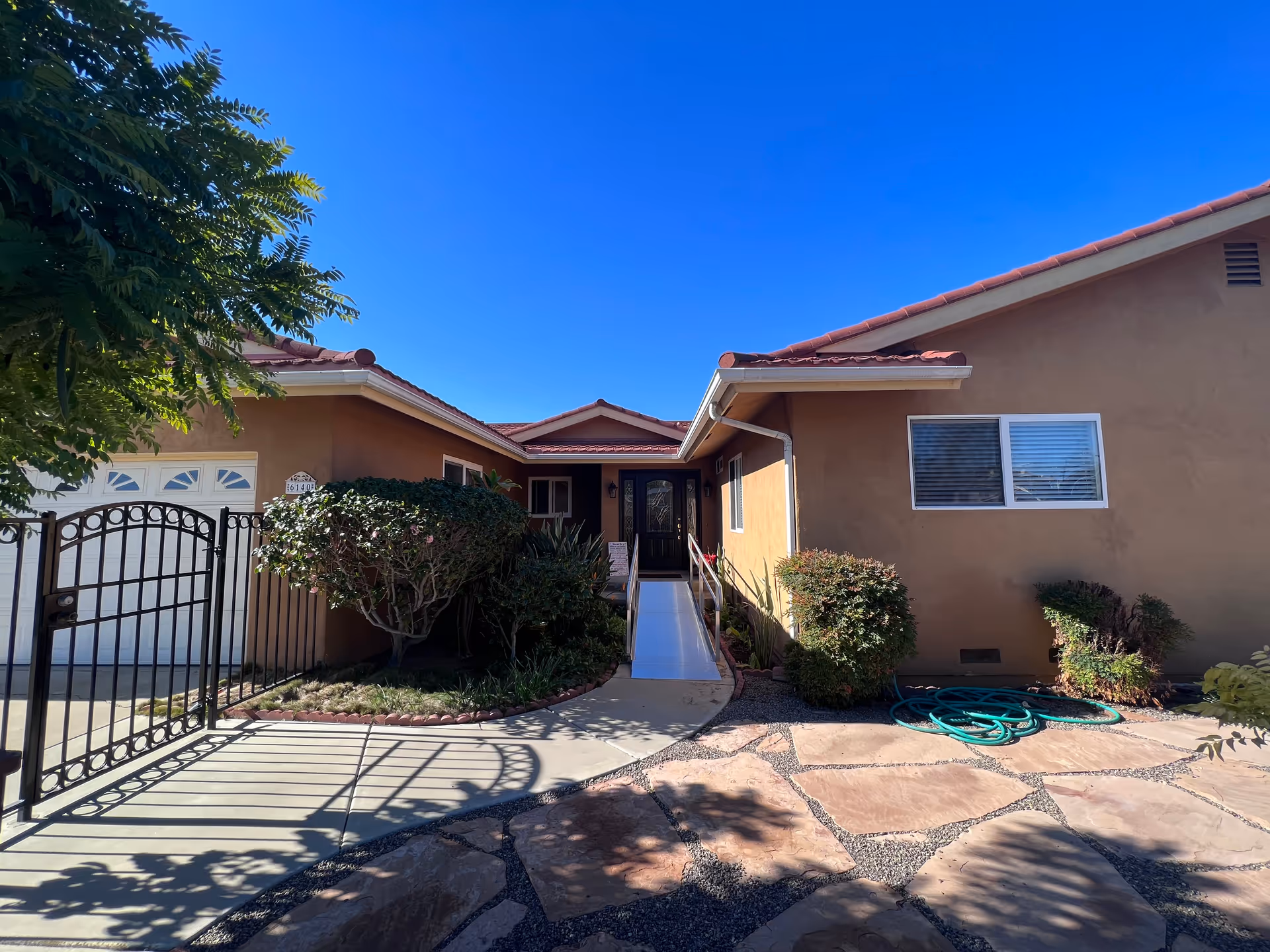 Front exterior of a single-story house with a gated driveway, a ramped entry, and landscaping under a clear blue sky.