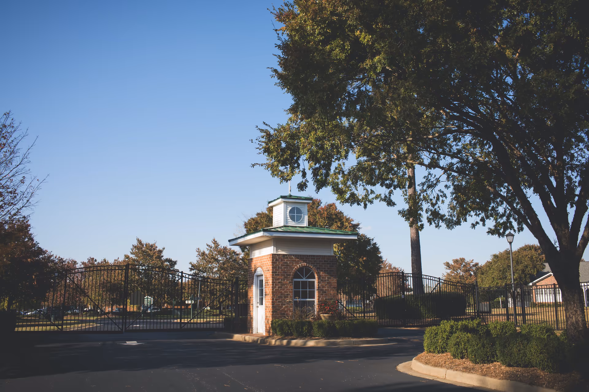 Gated entrance to a senior living facility with a small brick guardhouse topped with a green roof and a white cupola. The gate is black metal and surrounded by trees and landscaping under a clear blue sky.