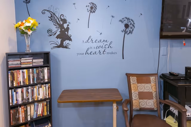 A small room with a blue wall decorated with a fairy and dandelion wall decals along with the quote 'A dream is a wish your heart makes'. There is a wooden table, a patterned cushioned chair, a black bookshelf filled with DVDs and books, a vase with yellow and white flowers on top of the bookshelf, and a mounted TV on the right side.