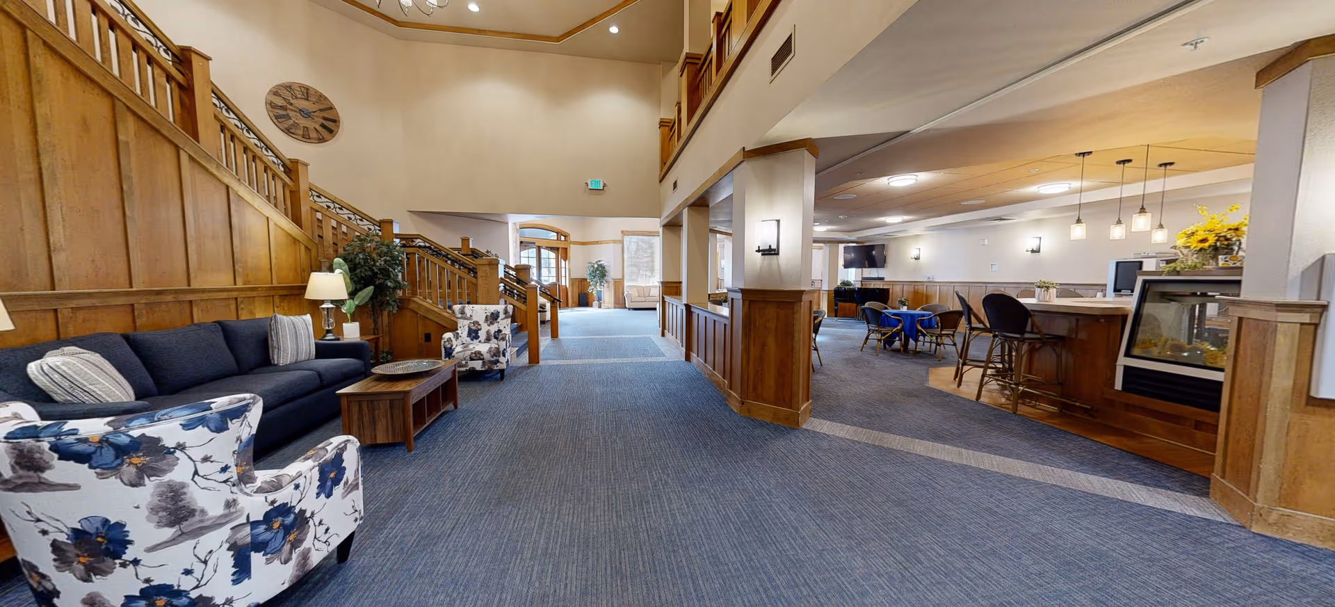 Spacious senior living facility common area with blue carpet, wooden paneling, and a staircase. The seating area includes a dark blue sofa, floral armchairs, and a wooden coffee table. To the right, there is a dining area with tables, chairs, a TV mounted on the wall, and pendant lights above a counter with bar stools. The space is well-lit with ceiling lights and natural light from windows.