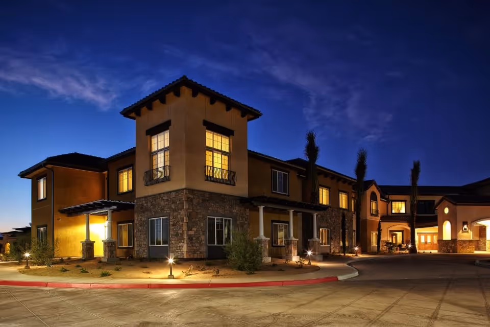Exterior view of Bella Villaggio senior living facility at dusk, showing a two-story building with warm interior lights, stone and stucco facade, and a curved driveway with small landscape lighting.