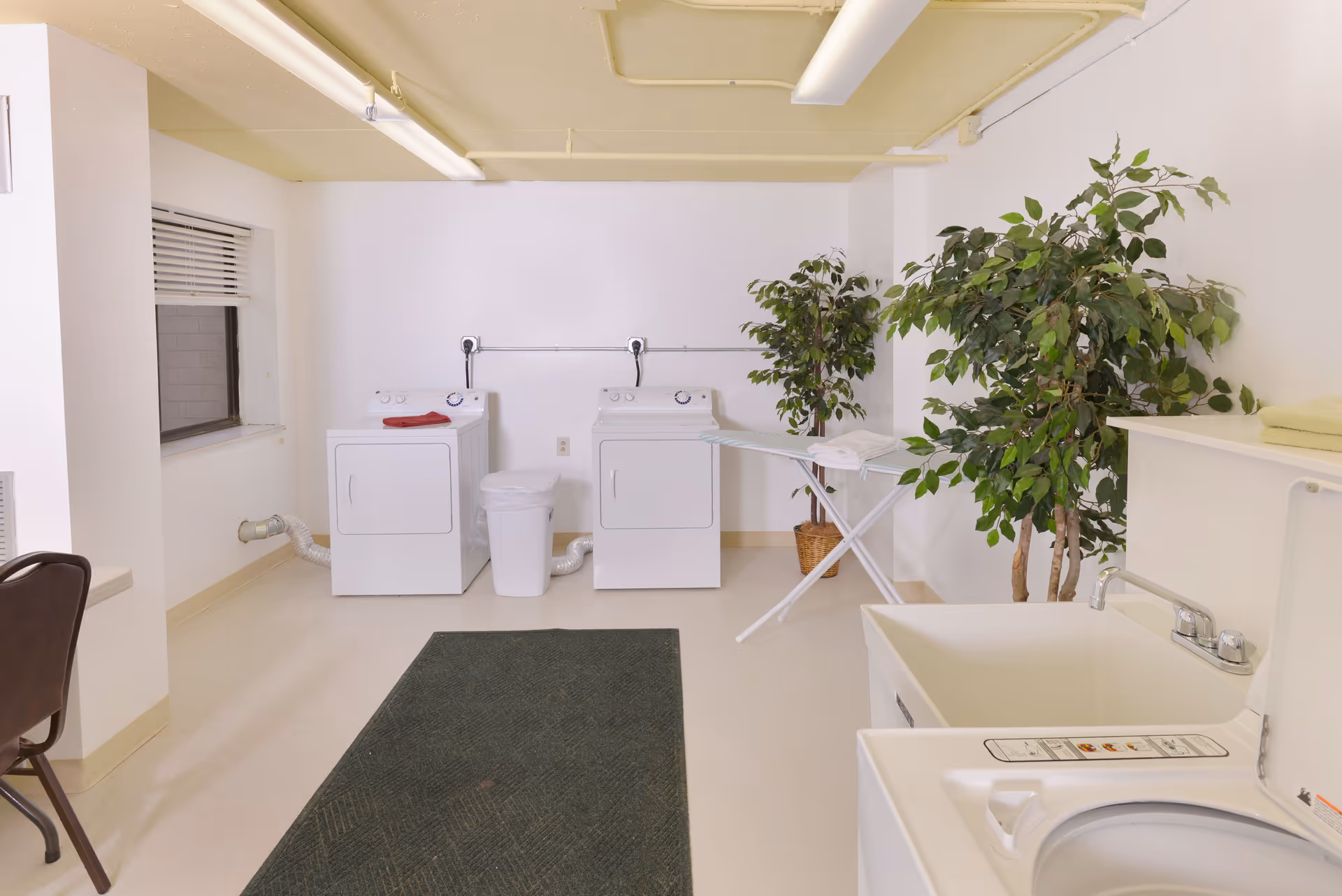 A clean laundry room with two white washing machines and a white trash bin between them. There is an ironing board with a white cloth on it, two large potted plants, a sink with a faucet, and a dark green floor mat. The room has white walls and a yellow ceiling with fluorescent lighting.