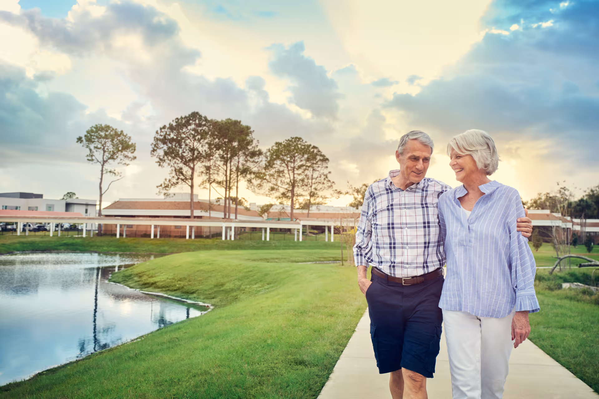 An elderly couple walking arm in arm along a paved path beside a pond with green grass and trees, with a building and a partly cloudy sky in the background during sunset.