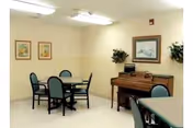 A small room with a round table and four chairs, a piano against the wall with a framed picture above it, two framed pictures on another wall, and two potted plants in the corners. The room has plain light-colored walls and ceiling lights.
