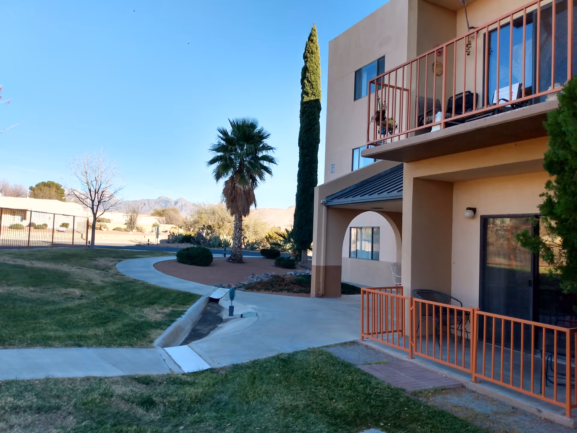 Exterior view of a beige multi-story senior living building with balconies overlooking a landscaped courtyard with palm trees, walkways, and distant mountains.