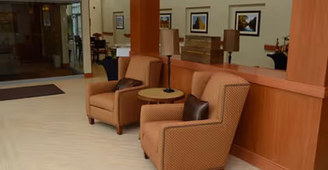 Two upholstered armchairs with a small round table and lamps in a senior living facility common area with wood paneling and framed artwork.