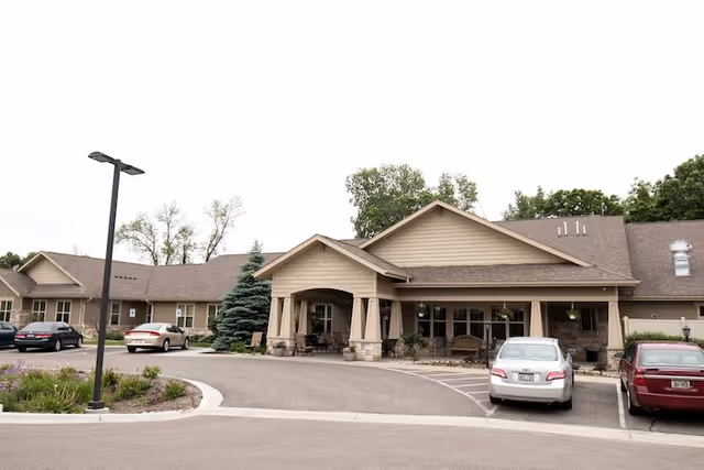 Exterior view of a single-story assisted living facility building with a covered entrance, several parked cars, a street lamp, and landscaping including trees and shrubs.