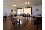 A spacious dining room with several tables covered with white tablecloths and chairs arranged around them. The room has wooden flooring, large windows with beige curtains, and ceiling lights. There is a crucifix on one wall and an air conditioning unit mounted on another wall.