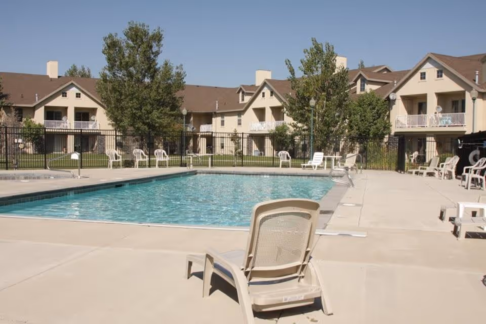 Outdoor swimming pool with lounge chairs in front of a multi-unit apartment building on a clear day.