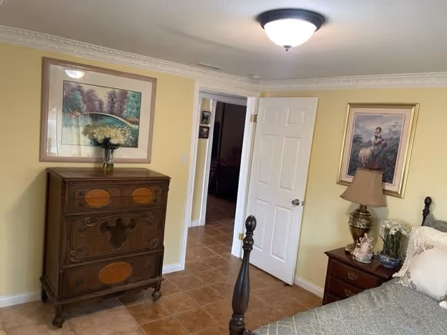 A bedroom with yellow walls, a wooden chest of drawers with a vase of flowers on top, an open white door leading to another room, a bedside table with a lamp and decorative items, and framed artwork on the walls.