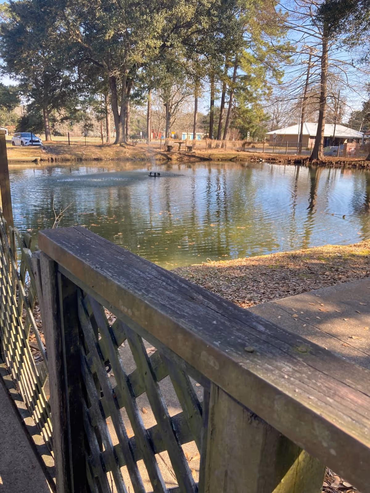 A peaceful outdoor scene featuring a small pond with a fountain in the center, surrounded by trees and a fenced area. A wooden railing and lattice fence are visible in the foreground, with a paved walkway beside the pond. The sky is clear and blue.