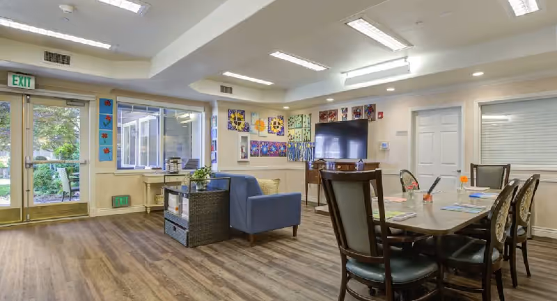 A well-lit common area in a senior living facility with wooden flooring, a blue armchair, a table with six chairs, colorful artwork on the walls, a large flat-screen TV, and glass doors leading outside to a garden area.