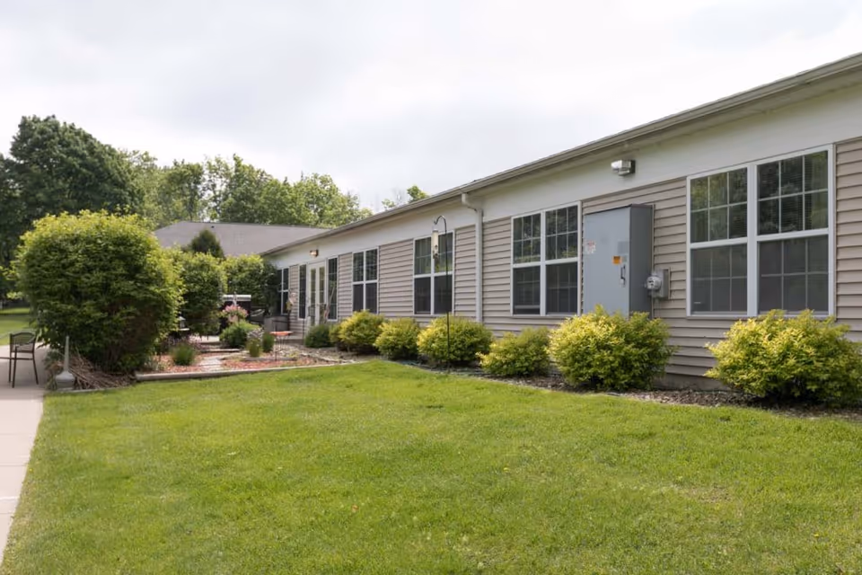 Single-story senior living building exterior with a grassy lawn, shrubs, and multiple large windows.