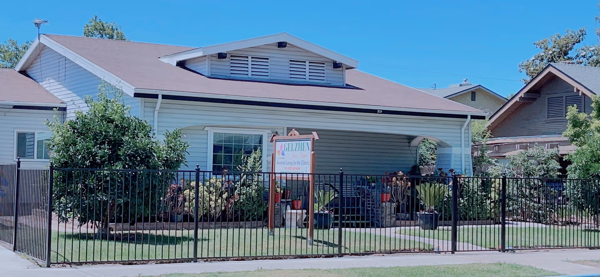 Exterior view of a single-story residential building with light gray siding and a brown roof. The front yard is enclosed by a black metal fence and features green grass, bushes, and potted plants. A sign in the yard reads 'Gelzhen Guest Home' indicating it is a residential living facility. The sky is clear and blue.