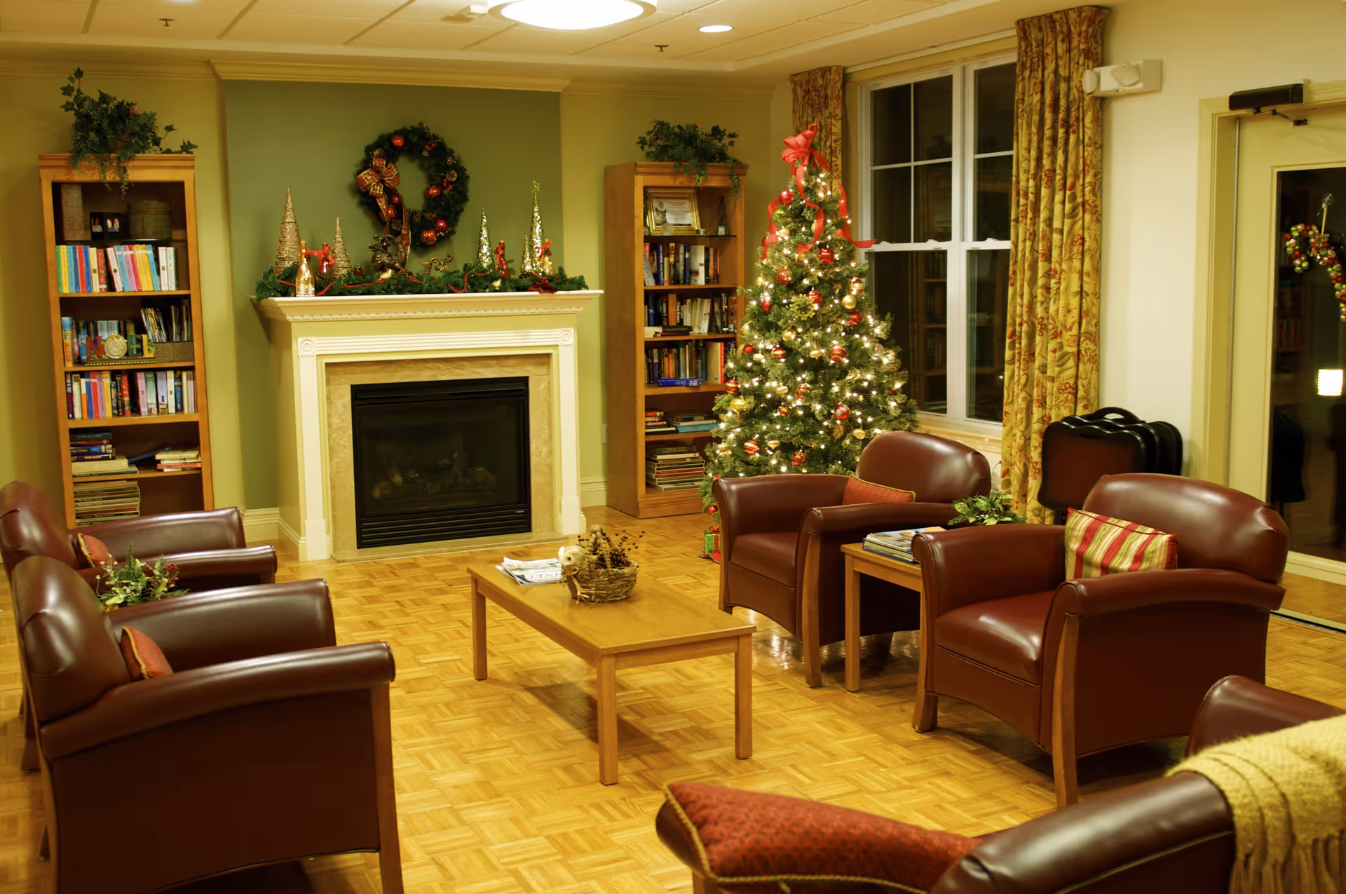 A cozy living room decorated for Christmas with a lit Christmas tree, a fireplace adorned with holiday decorations, two wooden bookshelves filled with books, and several brown leather armchairs arranged around a wooden coffee table on a parquet floor.