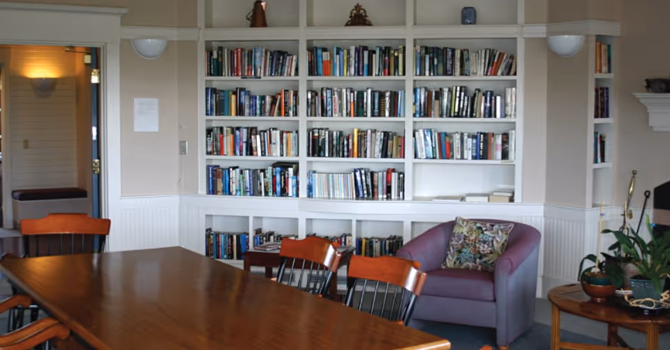 A cozy room with a large wooden table surrounded by chairs, a built-in white bookshelf filled with books, a purple armchair with a floral cushion, and a small round table with plants. There is a doorway leading to another room with a bench and soft lighting.