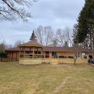 Single-story building with a wooden gazebo and ramp in front, surrounded by grass and leafless trees under a cloudy sky.
