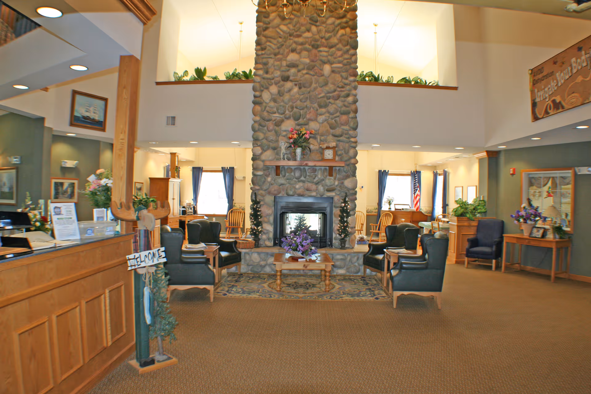 A spacious and warmly lit common area in a senior living facility featuring a large stone fireplace in the center with floral decorations on the mantle and a coffee table. Surrounding the fireplace are several comfortable armchairs arranged for conversation. The room has high ceilings with recessed lighting, wooden accents, and windows with blue curtains letting in natural light. There is a reception desk on the left with a welcome sign and various plants and framed pictures decorating the space.