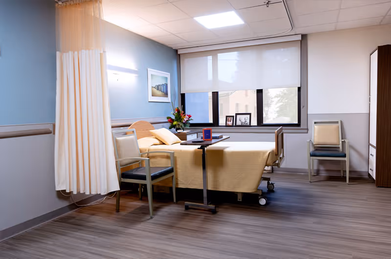 A bright rehabilitation patient room featuring a hospital bed with a beige cover, two chairs, a privacy curtain, and a window with framed photos.