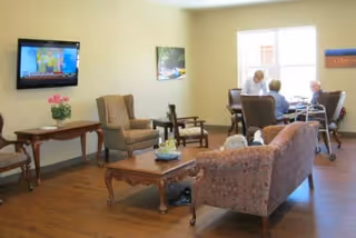 A cozy living room area in a senior living facility with wooden flooring, a floral patterned sofa, armchairs, a coffee table, and a wall-mounted television. Three elderly people are seated near a window, engaged in conversation or activity.