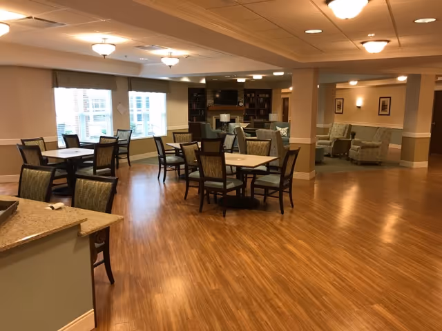 A spacious room with wooden flooring featuring several tables and chairs arranged for dining or socializing. The room has large windows with blinds, ceiling lights, and a cozy seating area with armchairs and a bookshelf in the background.