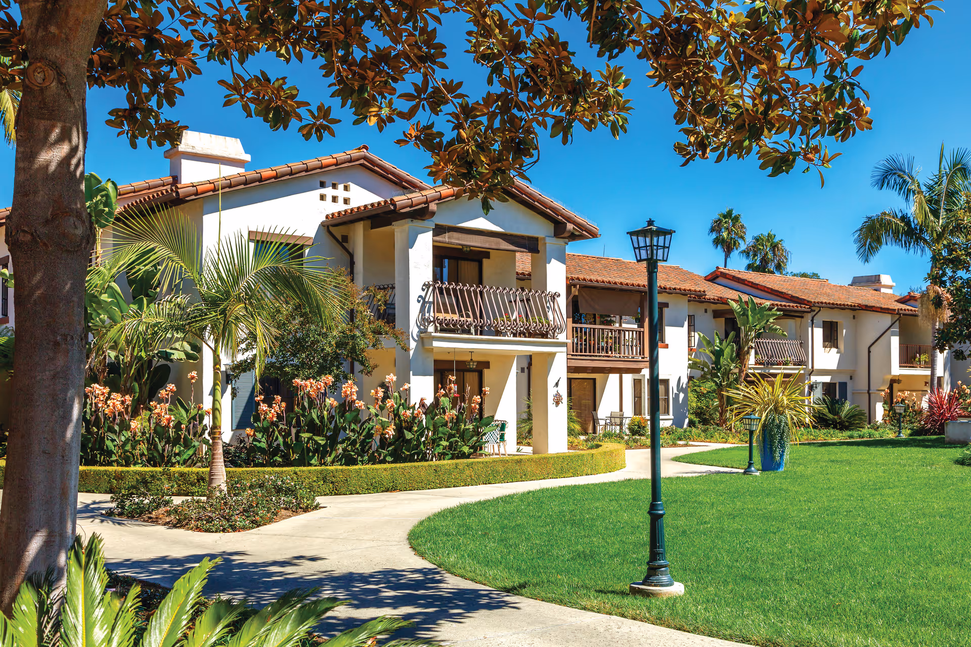 Exterior view of a senior living facility with white stucco buildings and red tile roofs surrounded by lush landscaping including palm trees, flowering plants, and green lawns under a clear blue sky. A paved walkway curves through the grass with vintage-style street lamps along the path.