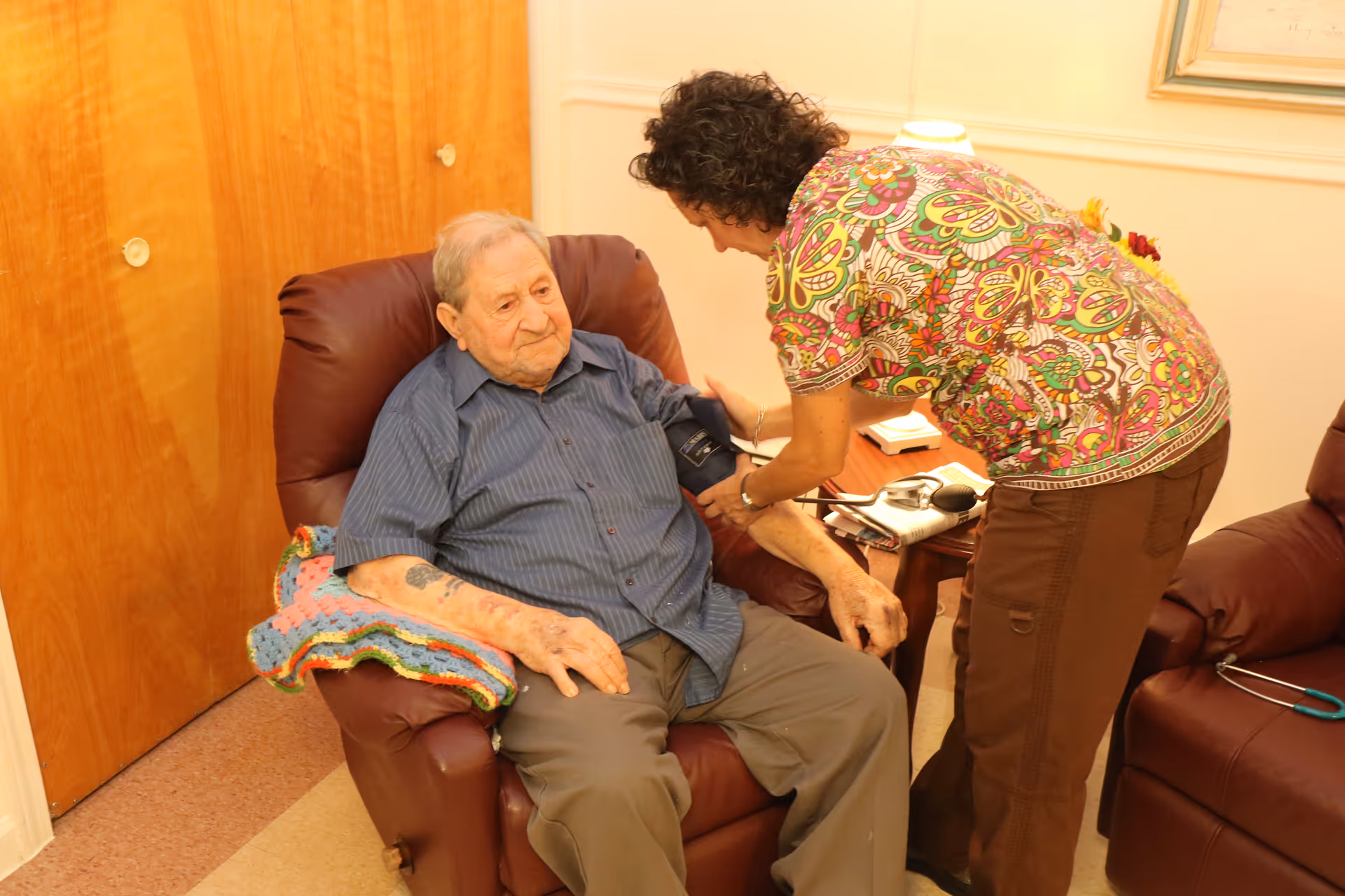 An elderly man sitting in a brown recliner chair while a healthcare worker in a colorful patterned shirt takes his blood pressure using a cuff on his arm. The setting appears to be a cozy room with wooden doors and a small table with medical equipment nearby.