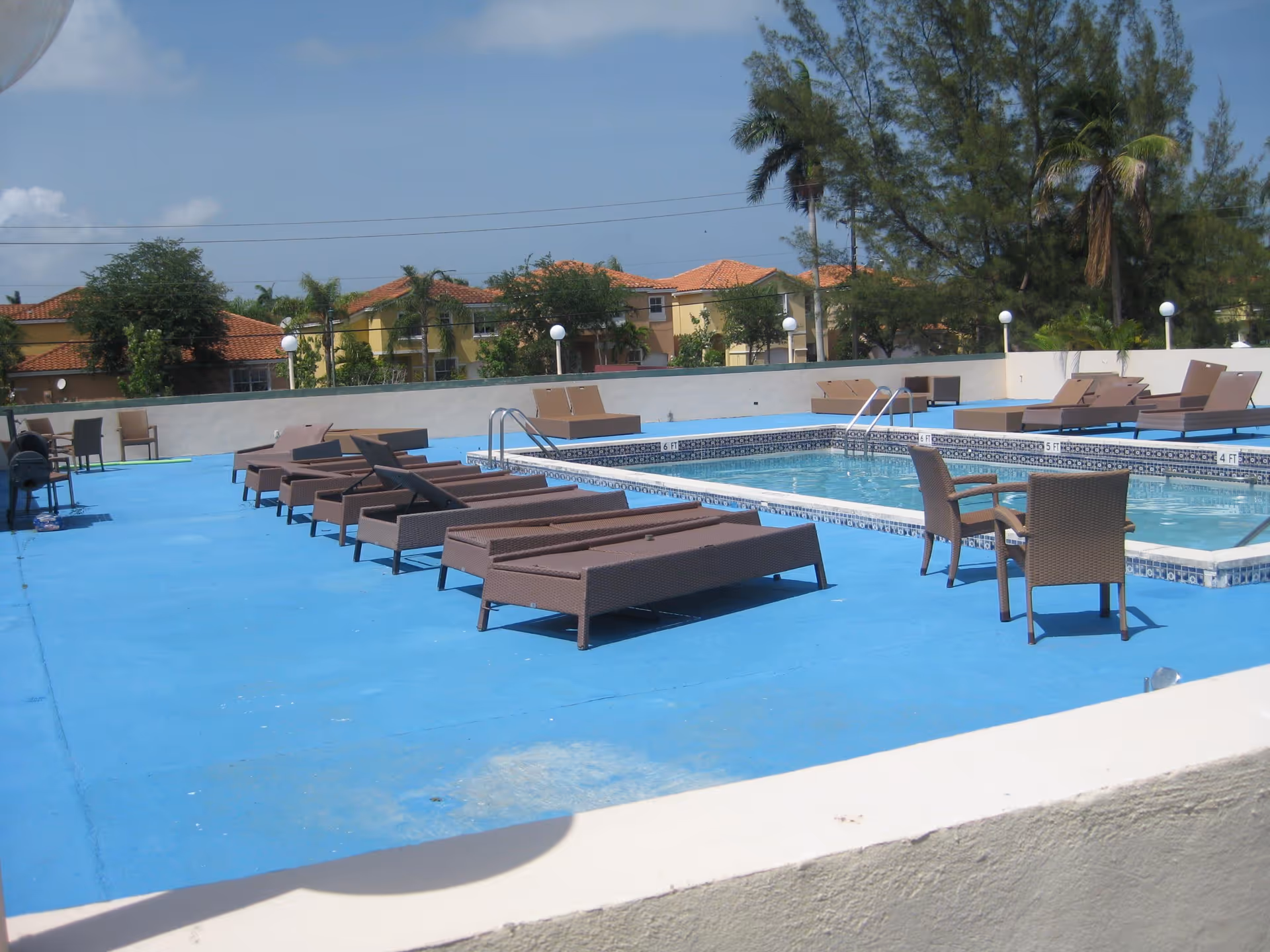 Outdoor swimming pool area with blue flooring, several brown lounge chairs and a couple of brown chairs with a small table. In the background, there are trees and houses with red-tiled roofs under a clear blue sky.