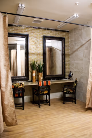 Interior view of a room with two large framed mirrors mounted on a tiled wall above a long counter. Two chairs with patterned cushions are placed in front of the counter. There are two potted plants on the counter, and beige curtains hang on either side of the room. The floor is wooden.