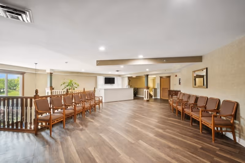 A spacious waiting area with two rows of wooden chairs with brown cushions facing each other. The room has wood flooring, beige walls, a large window showing green trees outside, a white reception desk with a mounted TV above it, and a mirror on the right wall.
