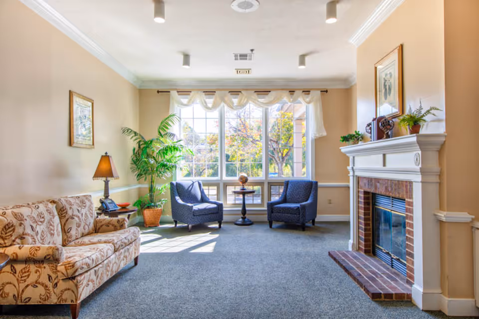 A bright and cozy living room with a large window letting in natural light. The room features a floral patterned sofa on the left, two blue armchairs near the window with a small round table between them, a potted plant, a table lamp, and a white fireplace with decorative items on the mantel.