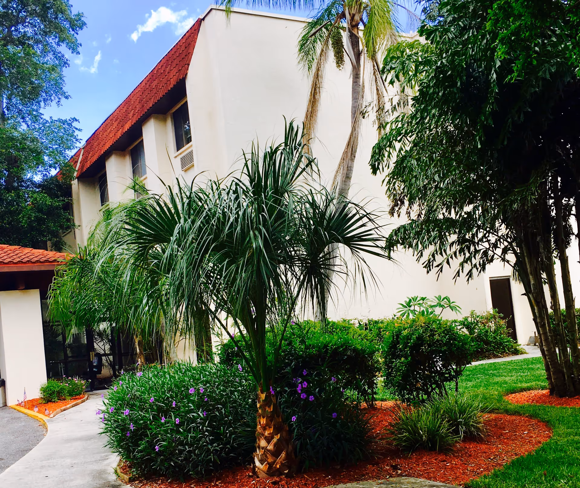 Exterior view of a building with a red-tiled roof and beige walls, surrounded by lush green landscaping including palm trees, bushes with purple flowers, and well-maintained grass under a partly cloudy blue sky.