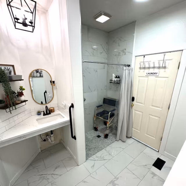 A clean, modern bathroom with white marble tile flooring and walls. The room features a walk-in shower with a shower chair and a curtain, a white countertop sink with a black faucet, a round mirror above the sink, and a door labeled 'LAUNDRY' with hooks holding a sign. There are also decorative plants on shelves next to the mirror and a black grab bar near the sink.