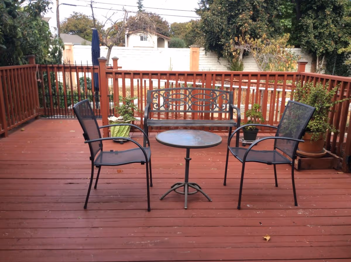 Wooden outdoor deck with two metal chairs, a small round table and a decorative bench beside potted plants and a red railing.