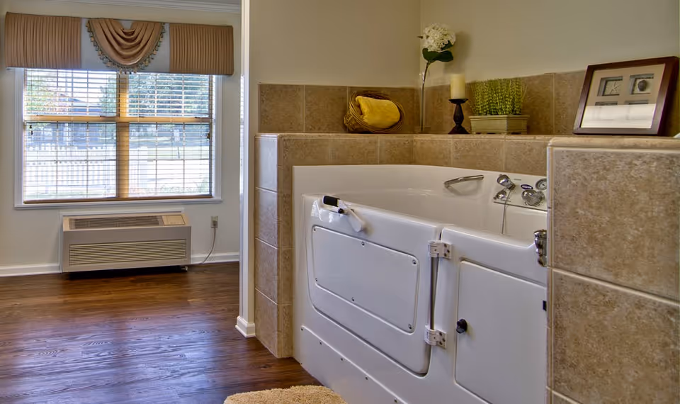 A bathroom featuring a walk-in bathtub with safety handles and a door for easy access. The tub is surrounded by beige tiles and decorated with a candle, a plant, a basket with towels, and a framed photo. There is a window with blinds and a valance, and a heating/cooling unit below the window. The floor is wooden with a small beige rug in front of the tub.