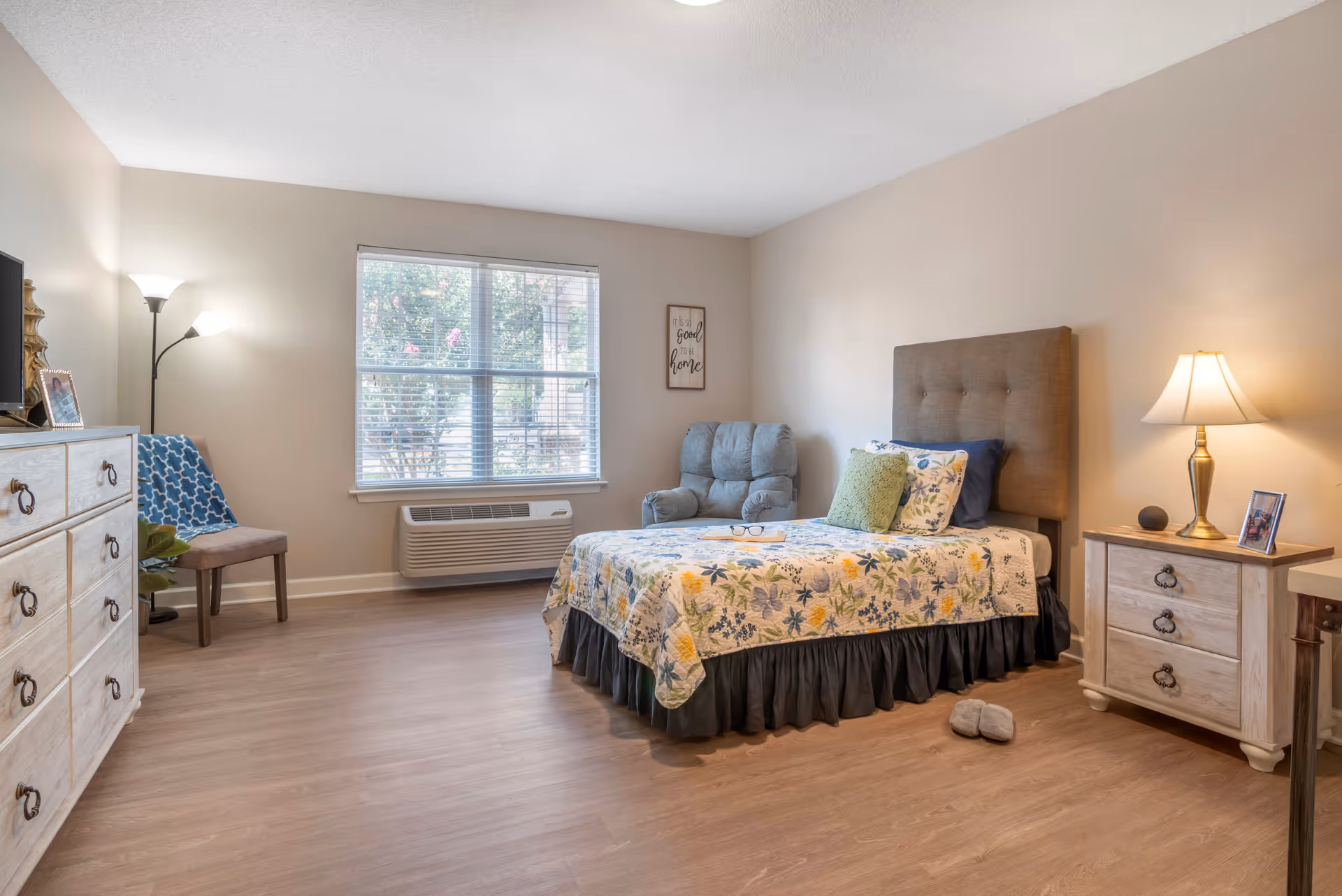 Bright furnished bedroom with a bed, armchair, dresser, nightstand, and window.
