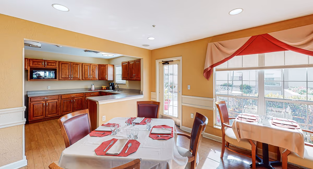 A bright dining area in a senior living facility with a table set for four, featuring red placemats, napkins, and glassware. The room has large windows with a red and beige valance, allowing natural light to fill the space. Adjacent to the dining area is a kitchen with wooden cabinets, a microwave, a coffee maker, and a sink. The walls are painted a warm beige color, and the floor is wooden.