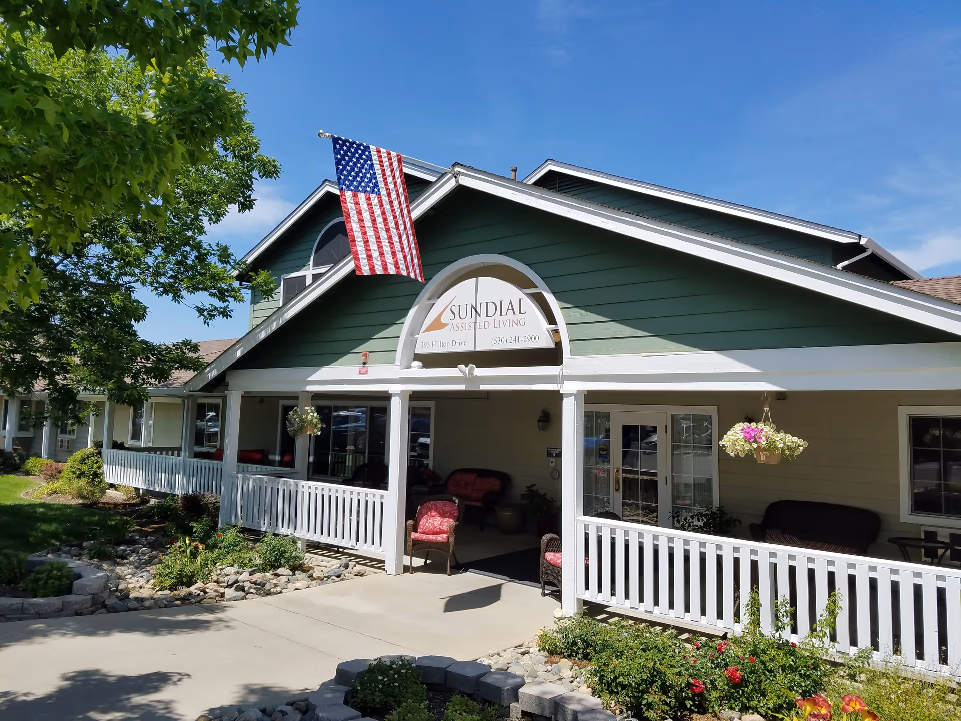 Exterior view of Sundial Assisted Living facility with a green and beige building, white railing, American flag hanging from the roof, and a garden with flowers and shrubs in front under a clear blue sky.
