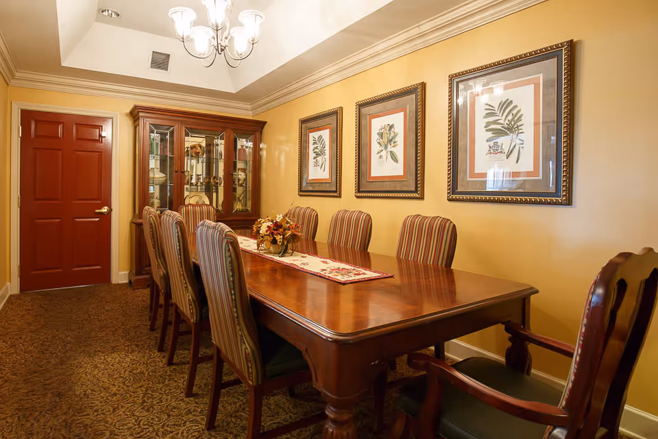 A formal dining room with a long wooden table surrounded by eight striped upholstered chairs. A floral centerpiece sits on a decorative table runner. The walls are painted yellow and adorned with three framed botanical prints. A wooden china cabinet with glass doors is positioned against the wall near a closed red door. A chandelier hangs from the ceiling, illuminating the room.
