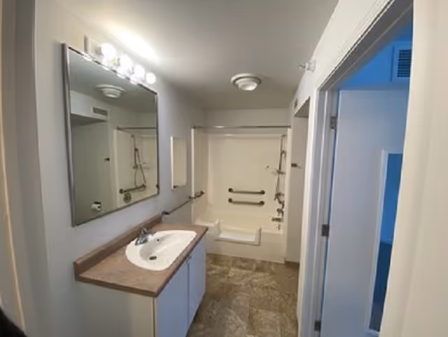 A clean bathroom with a large mirror above a sink with a countertop. The bathroom features a bathtub with grab bars and a built-in seat. The floor is tiled, and the walls are painted white. There is a door leading to another room on the right side.