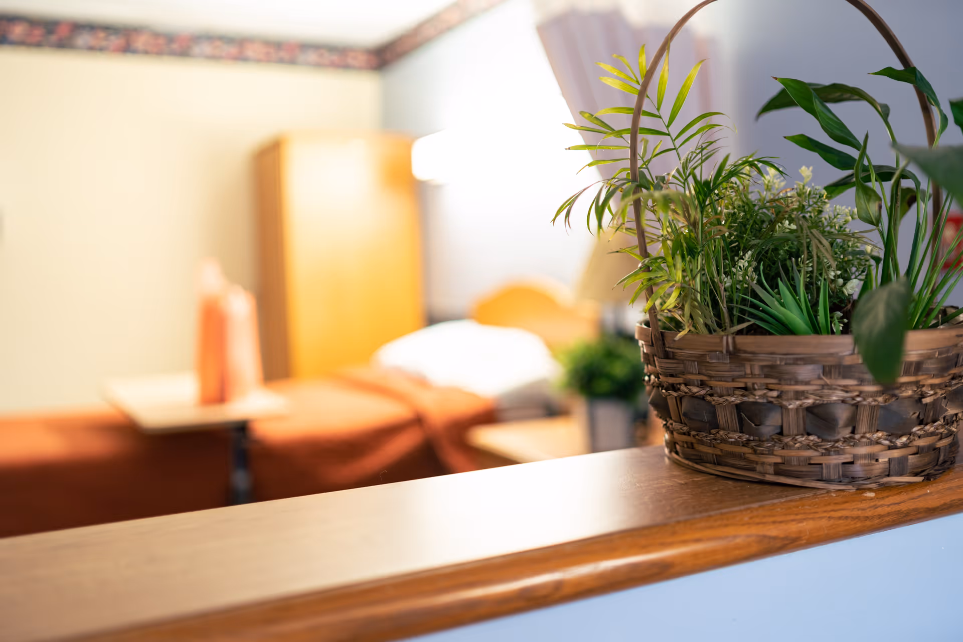 A close-up of a wicker basket filled with green plants on a wooden ledge, with a blurred background showing a bed with an orange blanket, a bedside table, and a lamp in a softly lit room.