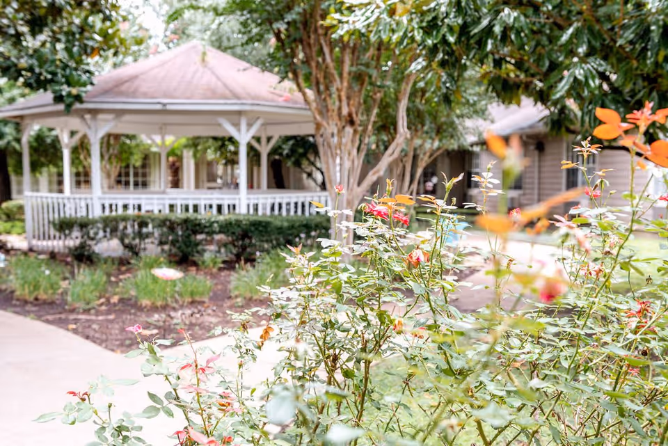 A garden area with flowering plants in the foreground and a white gazebo with a red roof in the background, surrounded by trees and shrubs at The Reserve at Pasadena Assisted Living and Memory Care.