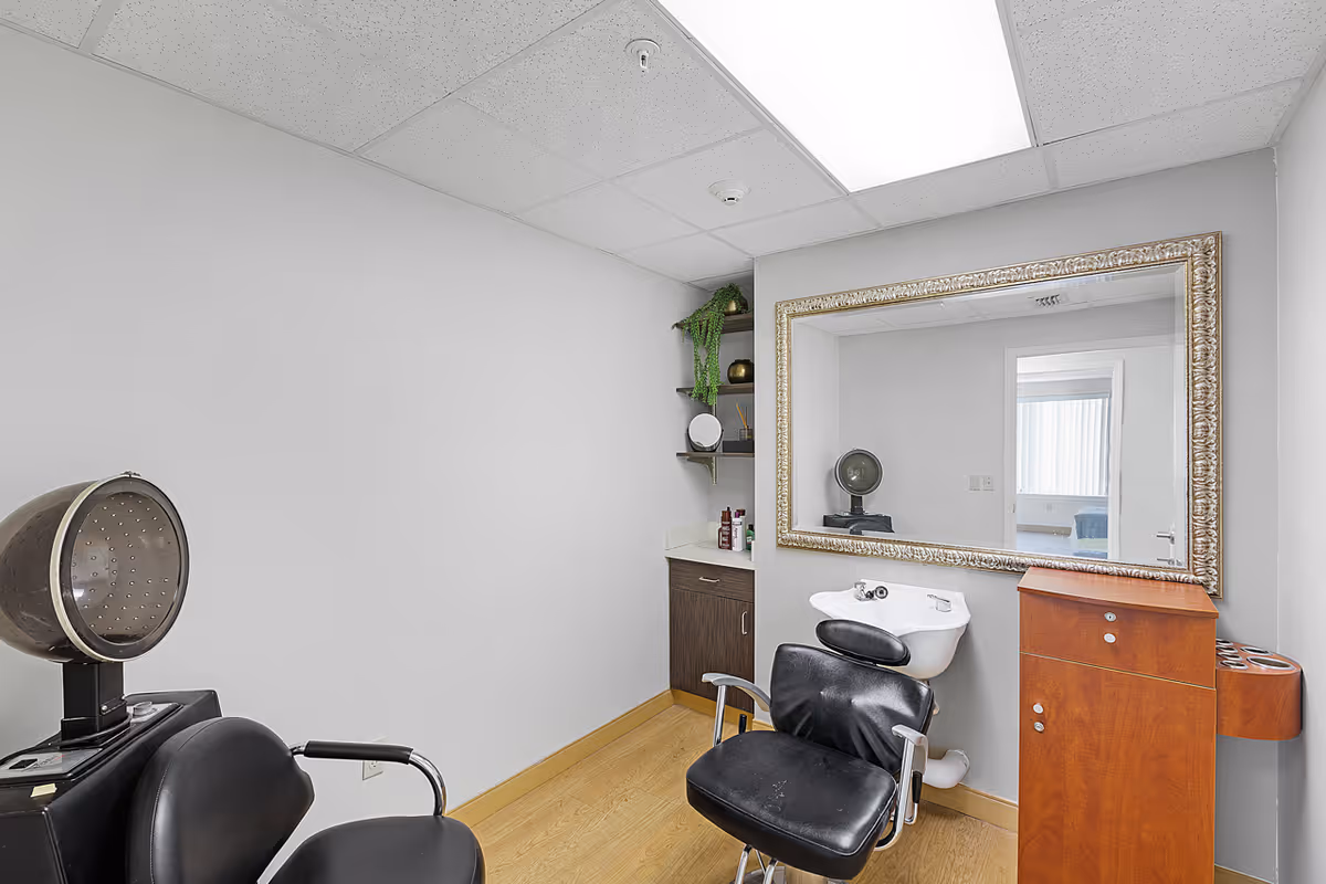 Interior of a small hair salon area with a black salon chair, a hair dryer hood, a wash basin, a large ornate mirror on the wall, wooden cabinetry, and shelves with decorative items and hair products.