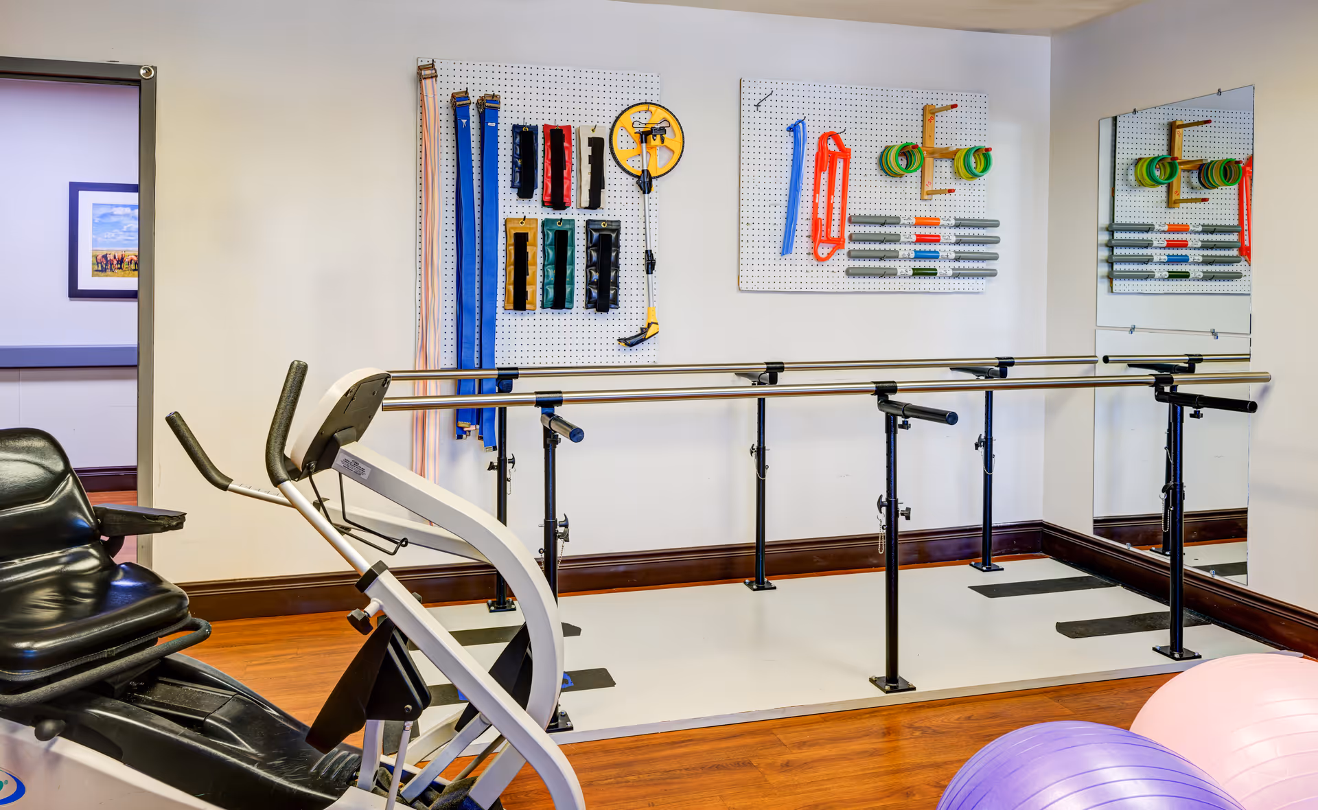 A physical therapy or exercise room with parallel bars for walking support, exercise bands and equipment hanging on pegboards on the wall, a recumbent exercise bike, a large mirror, and two exercise balls on a wooden floor.