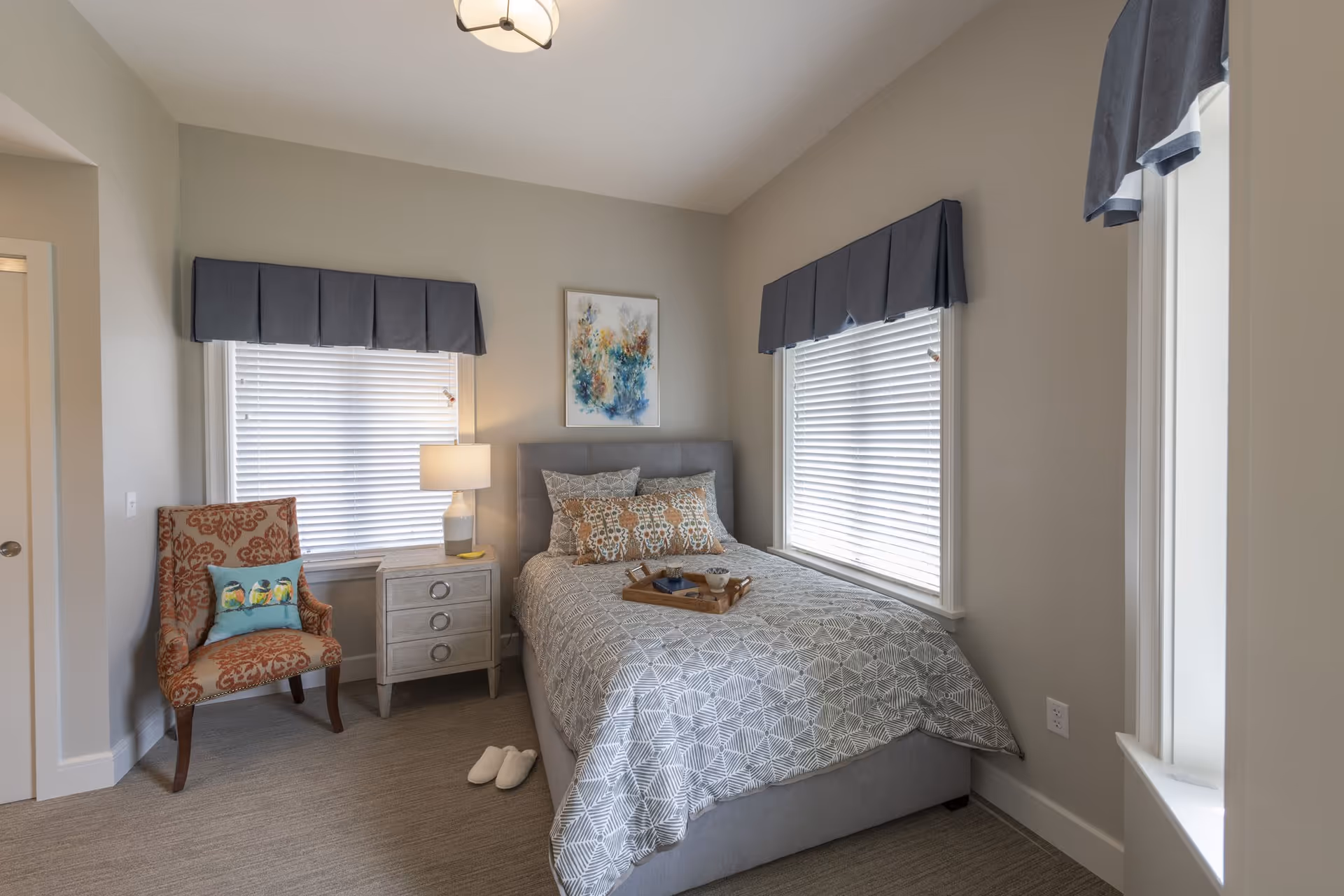 A cozy bedroom with a gray upholstered bed covered in a patterned gray and white comforter, decorative pillows, and a wooden tray with a cup and candle. There are two windows with white blinds and dark gray valances. A small white nightstand with a lamp is next to the bed, and an orange patterned armchair with a bird-themed pillow is placed near the window. The walls are painted light gray, and there is a colorful abstract painting above the bed. A pair of white slippers is on the carpeted floor.