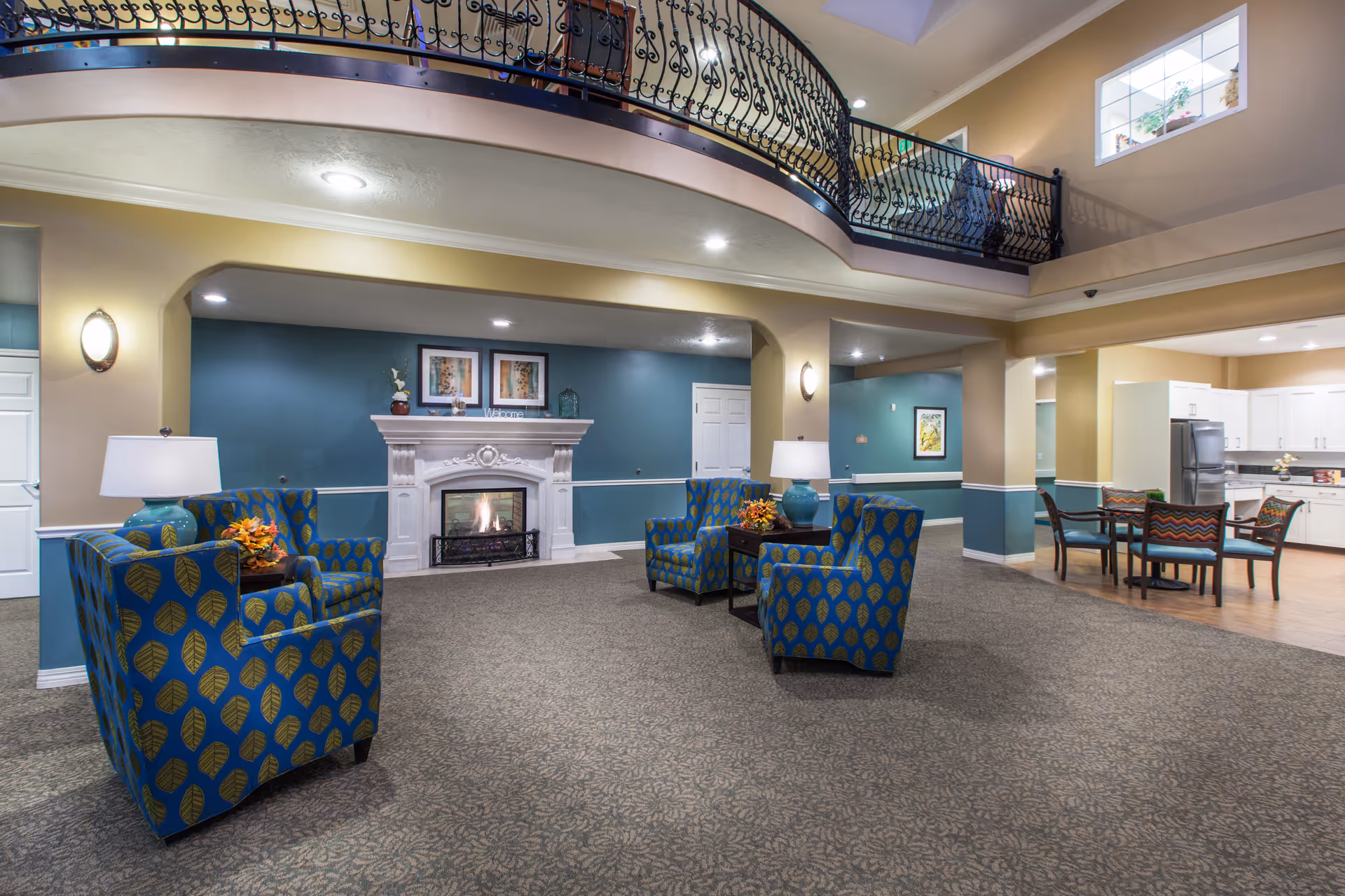 Open common lounge with patterned armchairs facing a fireplace, an overhead decorative balcony, and a dining/kitchen area to the right.