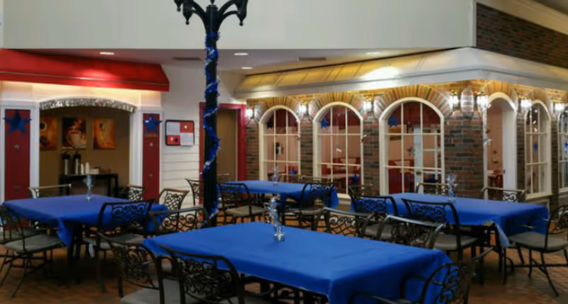 Indoor dining area with several tables covered in blue tablecloths and black wrought iron chairs. The space features a brick wall with arched windows and decorative lighting. A black lamp post wrapped with blue garland stands in the middle of the room.